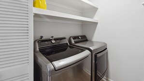 A laundry room with two stainless steel appliances, a white shelving unit above, and a yellow bag on the shelf. - Bright 3BR/2BA Home  Furnished w/yard + All Utilities Included near Lake Houston (Humble)