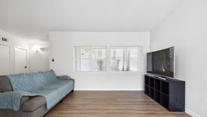 A room with a grey sofa, a TV on a black stand, and two windows with blinds. - Bright 3BR/2BA Home  Furnished w/yard + All Utilities Included near Lake Houston (Humble)