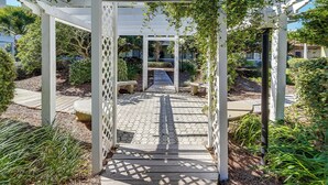 Une pergola en bois, ornée d’un motif en treillis, est entourée d’un jardin où poussent différentes espèces de plantes, et un chemin pavé traverse ce jardin.