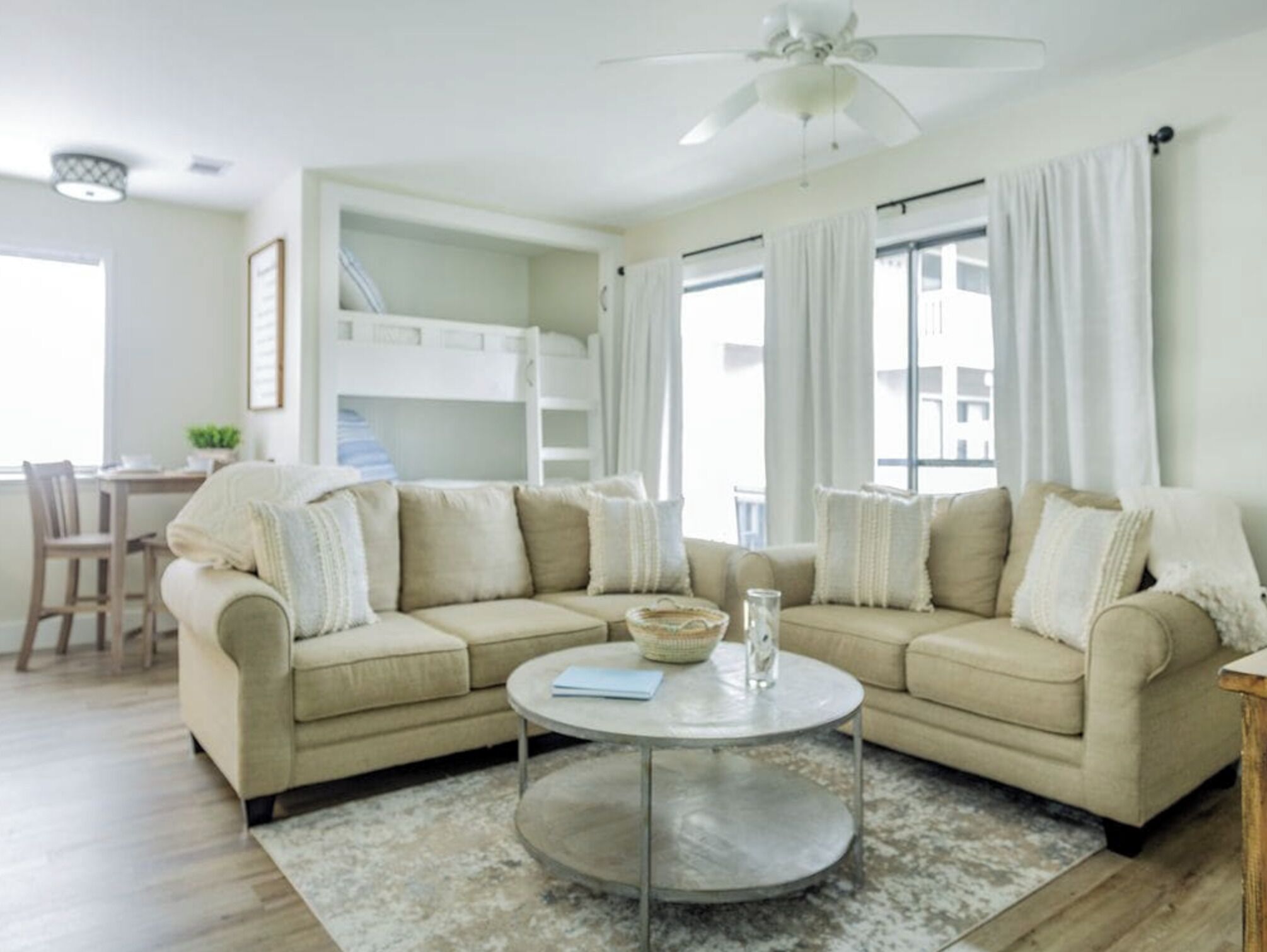 Una sala de estar con un sofá seccional color beige, una mesa redonda y una cama litera al fondo.