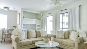 Un salon avec un canapé d’angle beige, une table basse ronde et, au fond, un lit superposé.
