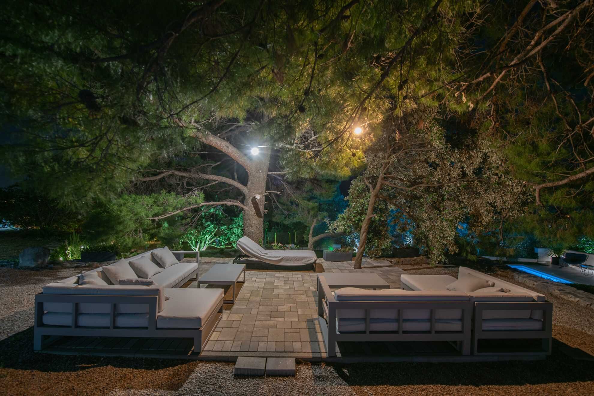 Un espacio al aire libre con sillones de descanso y una chimenea, rodeado de árboles e iluminado con luces ambientales.