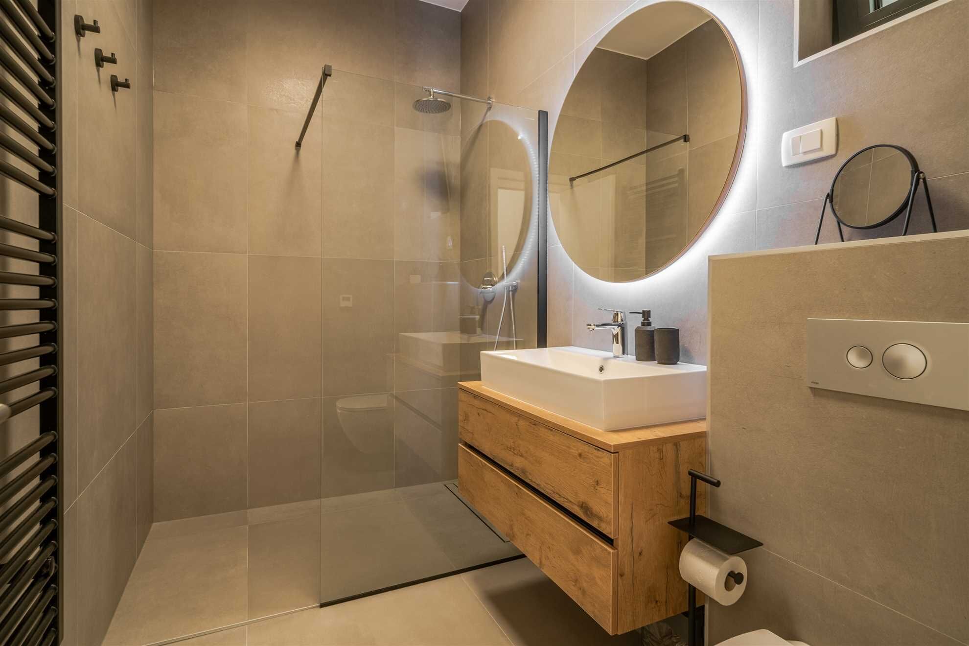 Un baño moderno con ducha envuelta en vidrio, tocador de madera con lavamanos y dos espejos redondos.