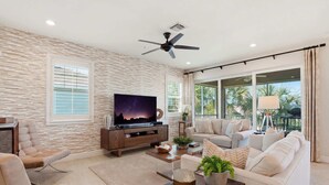 A modern living room with a flat-screen TV, a ceiling fan, a sofa, a coffee table, and a large window with blinds. - Lakefront Luxury + Stunning Naples Sunsets (Naples)