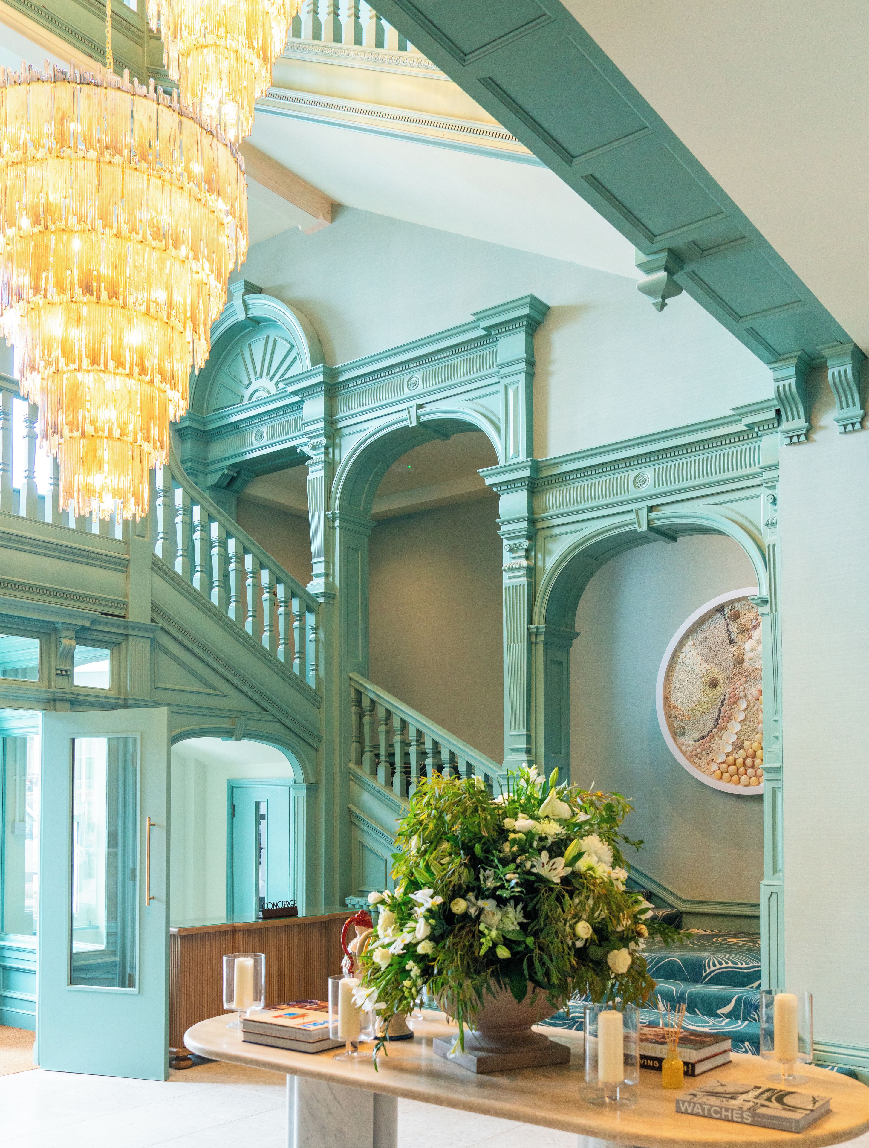 A grand staircase with ornate blue detailing, a chandelier, and a table with a floral arrangement and books.