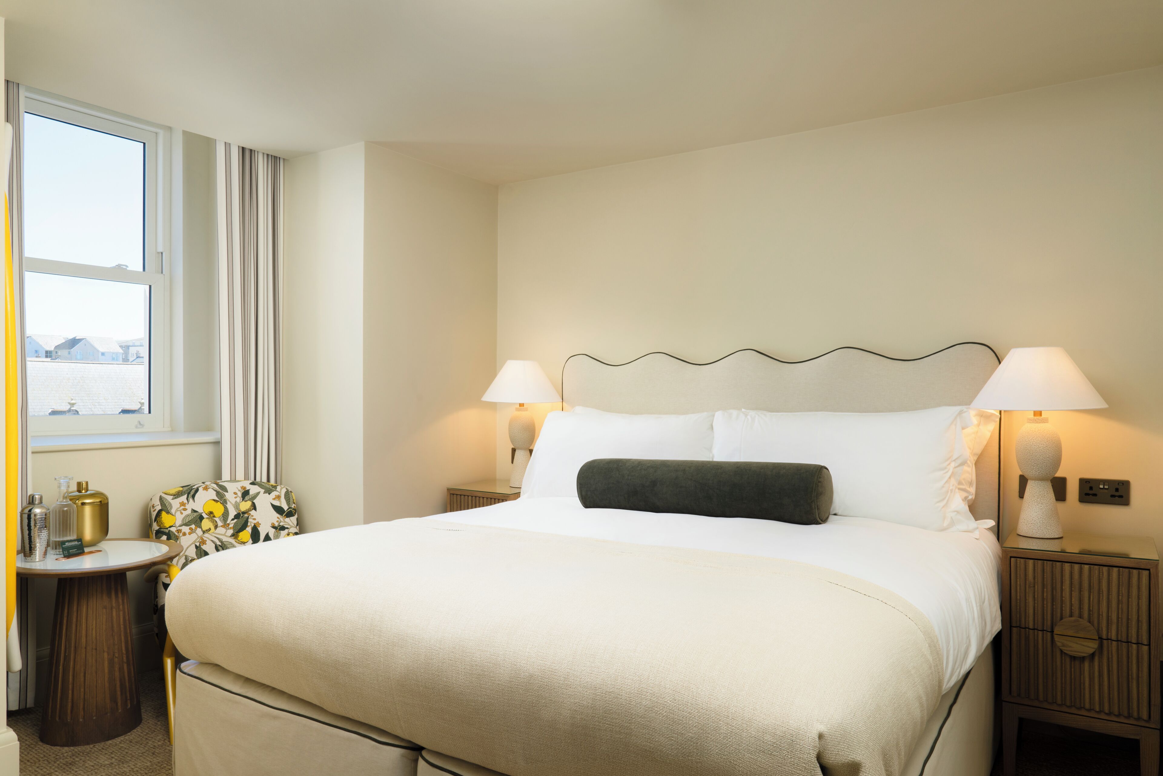 A hotel room with a large bed, two bedside lamps, a nightstand with a lamp, a small table with a lamp, and a window with curtains.