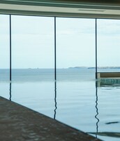 Vista a través de amplios ventanales de una piscina con un mar tranquilo y tierra al fondo.