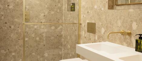 Un baño moderno con ducha envuelta en cristal, un lavabo blanco y suelo de mármol.