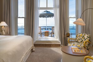 A hotel room with a bed, a round table, a chair with a floral pattern, a balcony with a table and chairs, and a view of the sea.