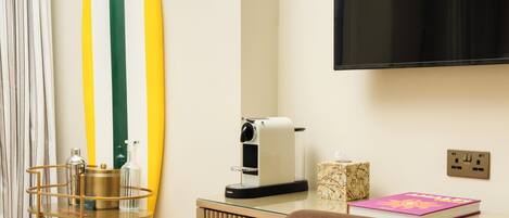 A room with a wooden desk, a television mounted on the wall, a coffee machine, a surfboard decoration, and a chair.