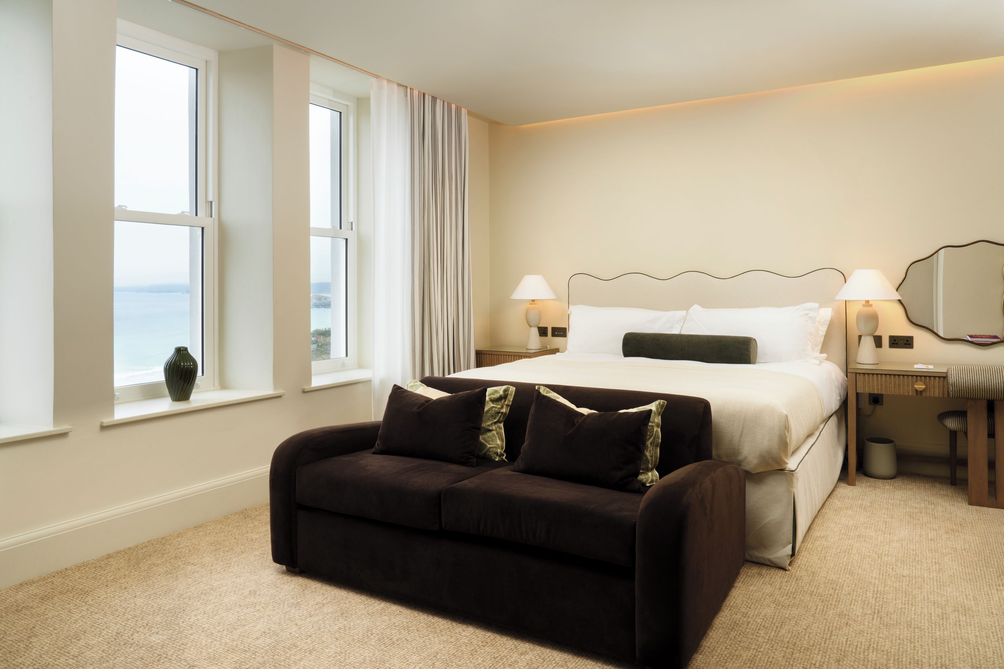 A hotel room with a bed, bedside lamps, a sofa, a window with curtains, and a view of the sea.