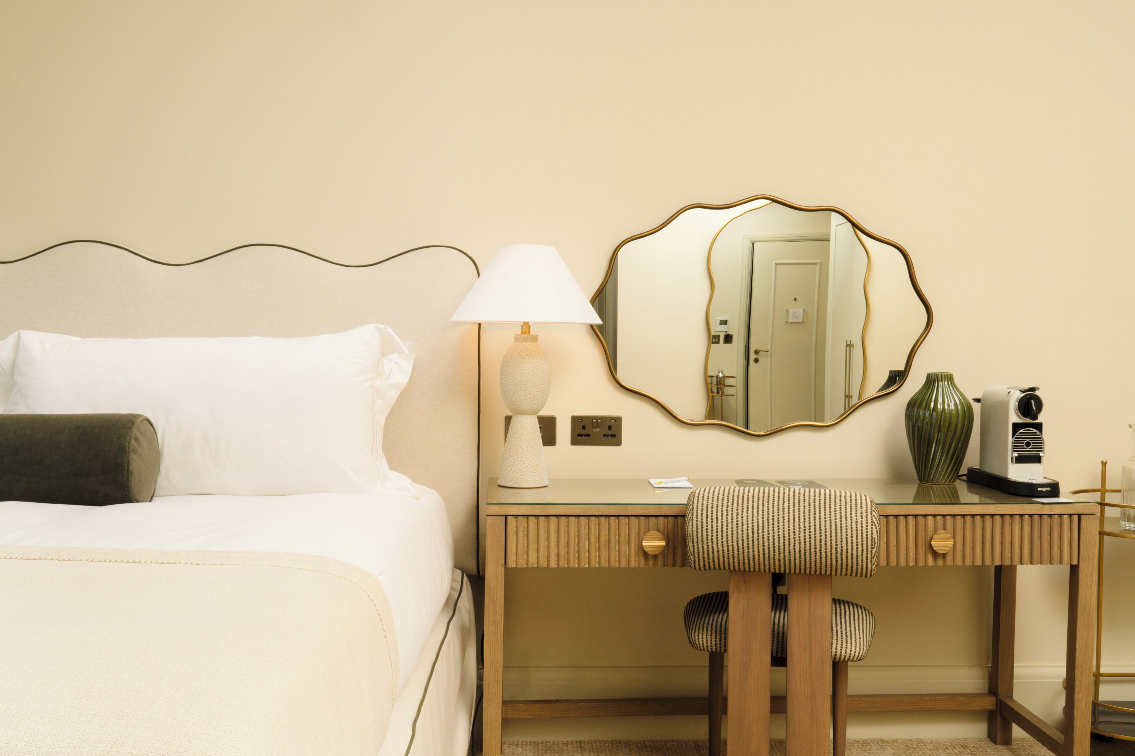 A hotel room with a bed, a desk, a chair, a lamp, a mirror, and a vase.