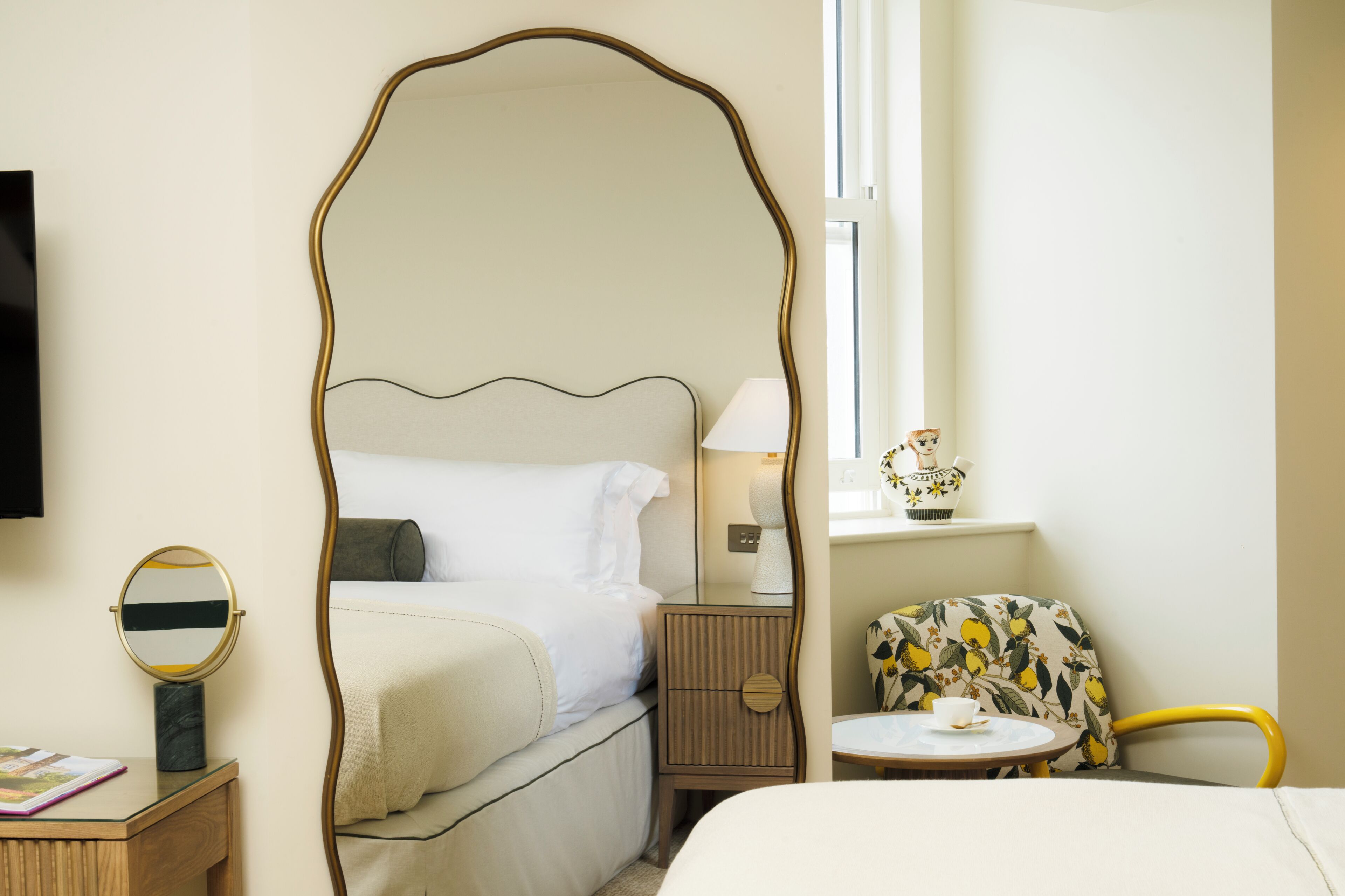 A bedroom with a four-poster bed, a bedside table with a lamp, a chair with a floral pattern, and a small table with a cup.
