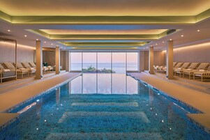 Una amplia piscina interior con agua cristalina, rodeada de sillones de descanso y vistas al mar.