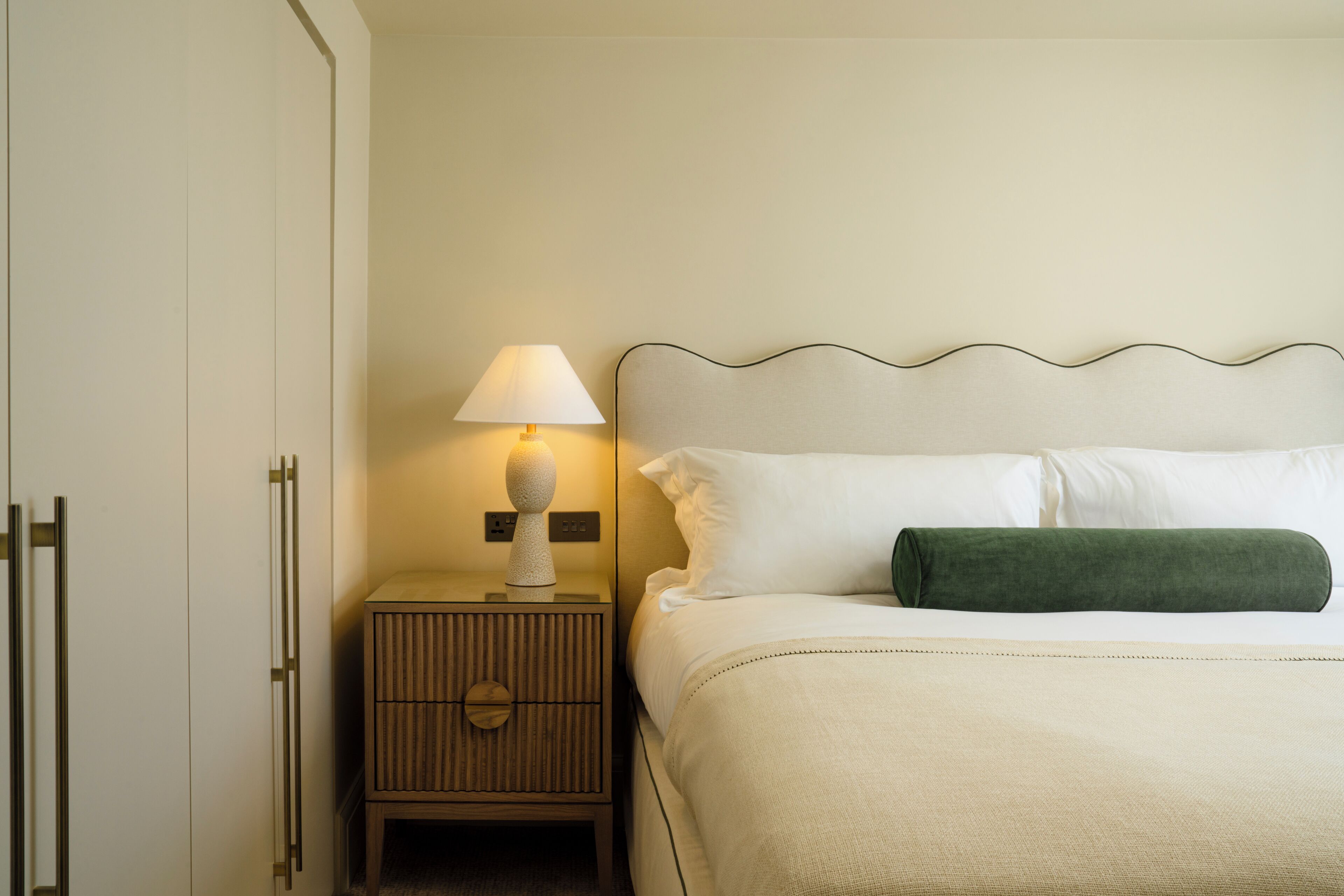 A neatly made bed with white linens and a green pillow, a bedside lamp, and a wooden nightstand with a lamp.