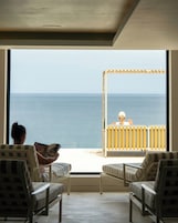 Um quarto com uma grande janela com vista para o mar, duas cadeiras e uma pessoa lendo um livro.