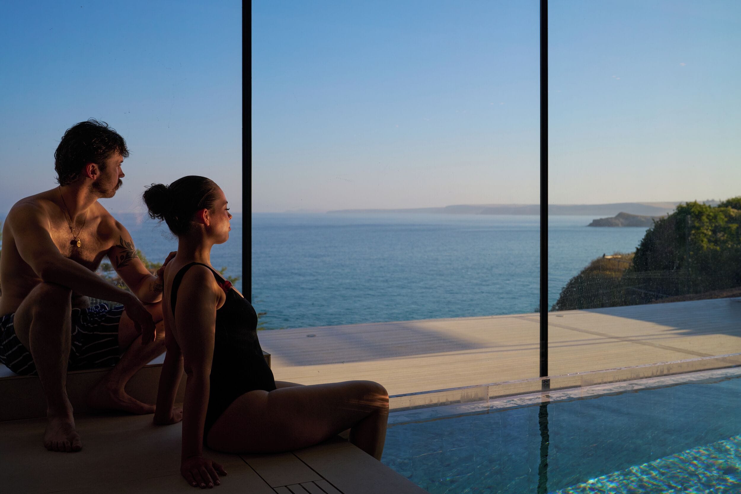 A couple sitting by a pool with a view of the sea and a cliff.