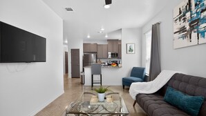Ein modernes Wohnzimmer mit Glastisch, Sofa, Flachbildfernseher und einer Küche im Hintergrund.