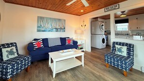 A cozy living room with a blue sofa, matching armchairs, a white coffee table, and a ceiling fan. A painting of sailboats hangs on the wall, and a kitchen area is visible in the background. - The Tinker Bell at Moody Beach- Turtle Unit 2 BR ’s with partial ocean views (Wells)