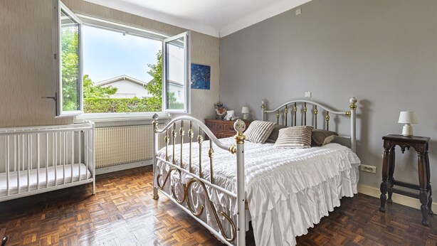Una camera da letto con un letto in ferro bianco, una panca di legno, una finestra vista e un tavolino con una lampada.