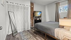 A bedroom with a bed, a TV, a brick wall, and a window with curtains.