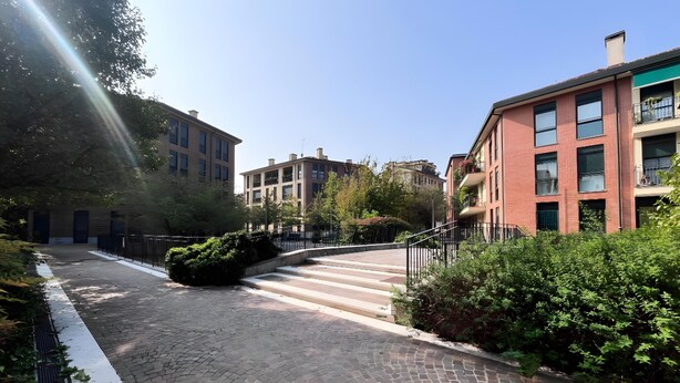 Ein gepflasterter Weg führt zu einem Gebäude mit einer Treppe, umgeben von Grünflächen und weiteren Gebäuden.