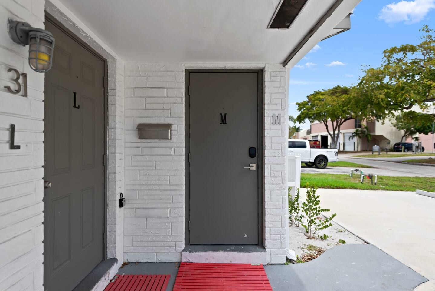 A residential entrance with a white brick facade, two doors labeled 'L' and 'M', and a red mat at the entrance.