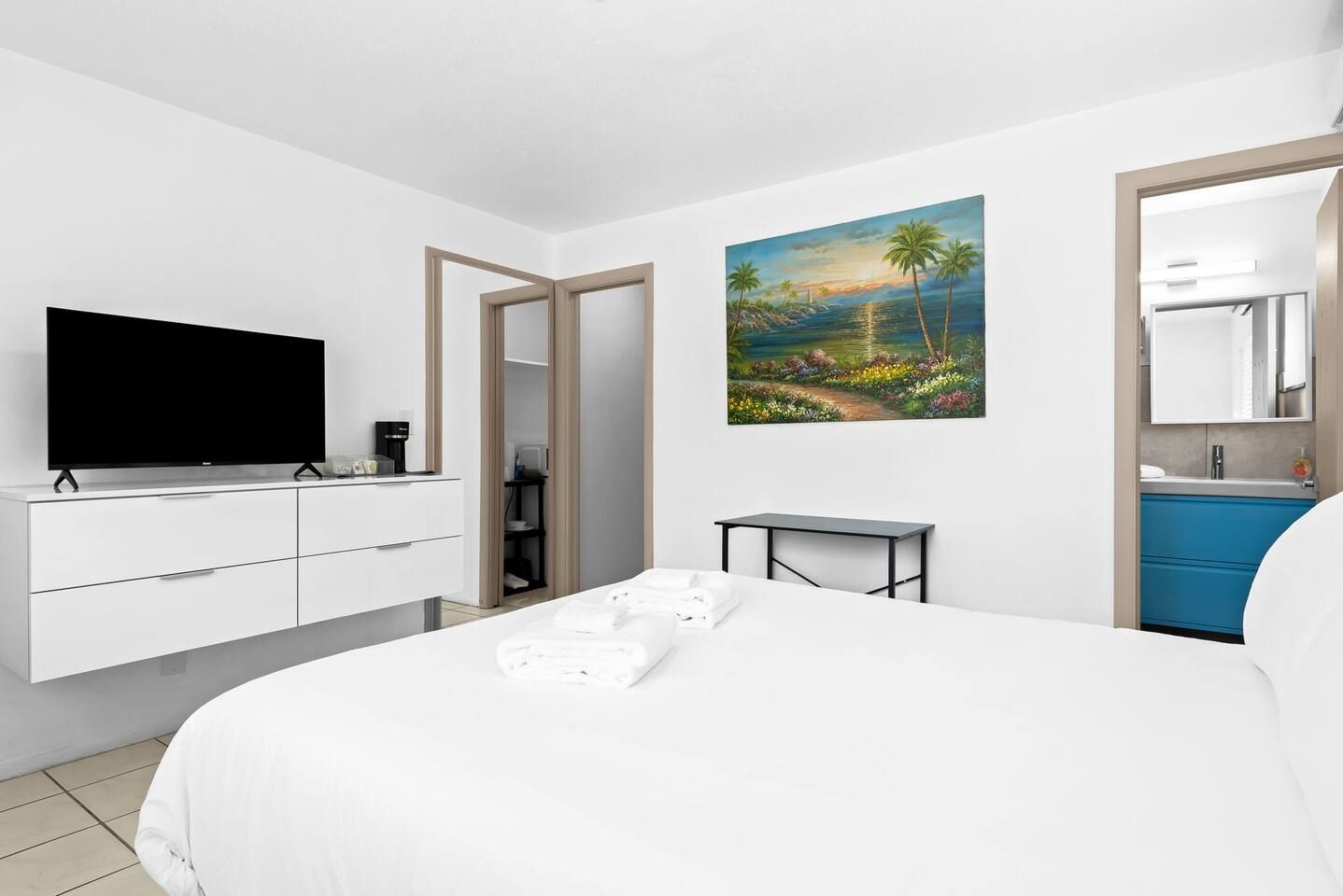 A hotel room with a bed, a TV, a painting, a desk, and a bathroom.