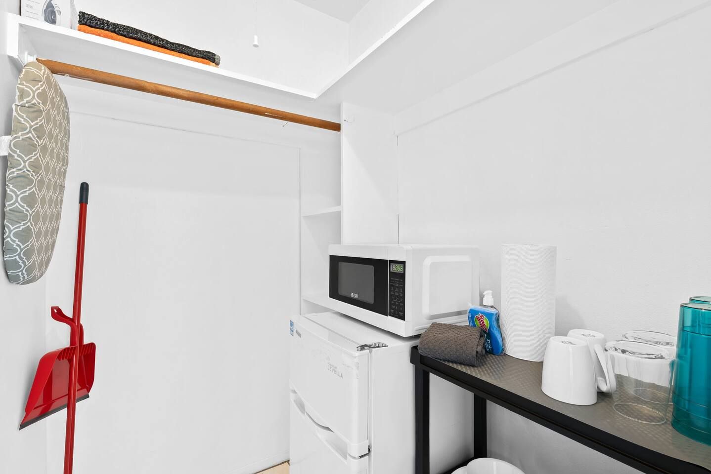 A compact kitchen with a microwave, a kettle, and a small table with cups and a bottle.