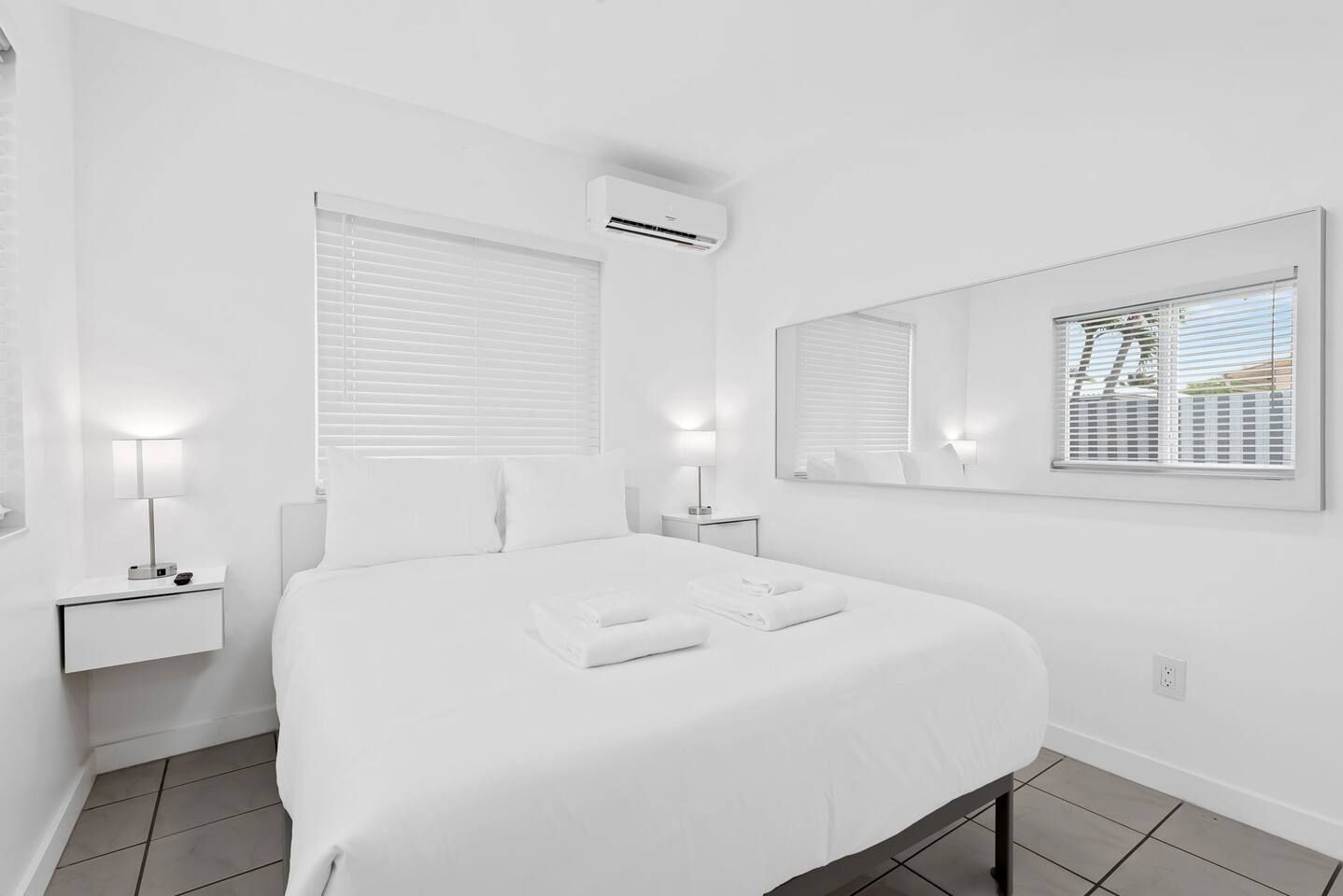 Une chambre d’hôtel avec un grand lit, deux tables de chevet avec des lampes, un climatiseur fixé au mur et une fenêtre avec des stores.