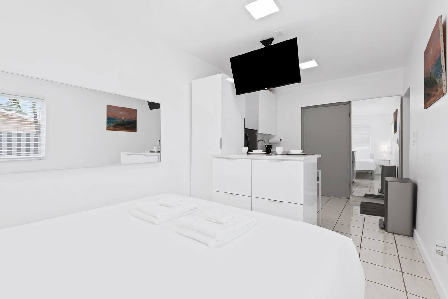 Une chambre d’hôtel moderne dotée d’un grand lit, d’un téléviseur à écran plat, d’une petite kitchenette et d’une salle de bain visible à travers une porte ouverte.