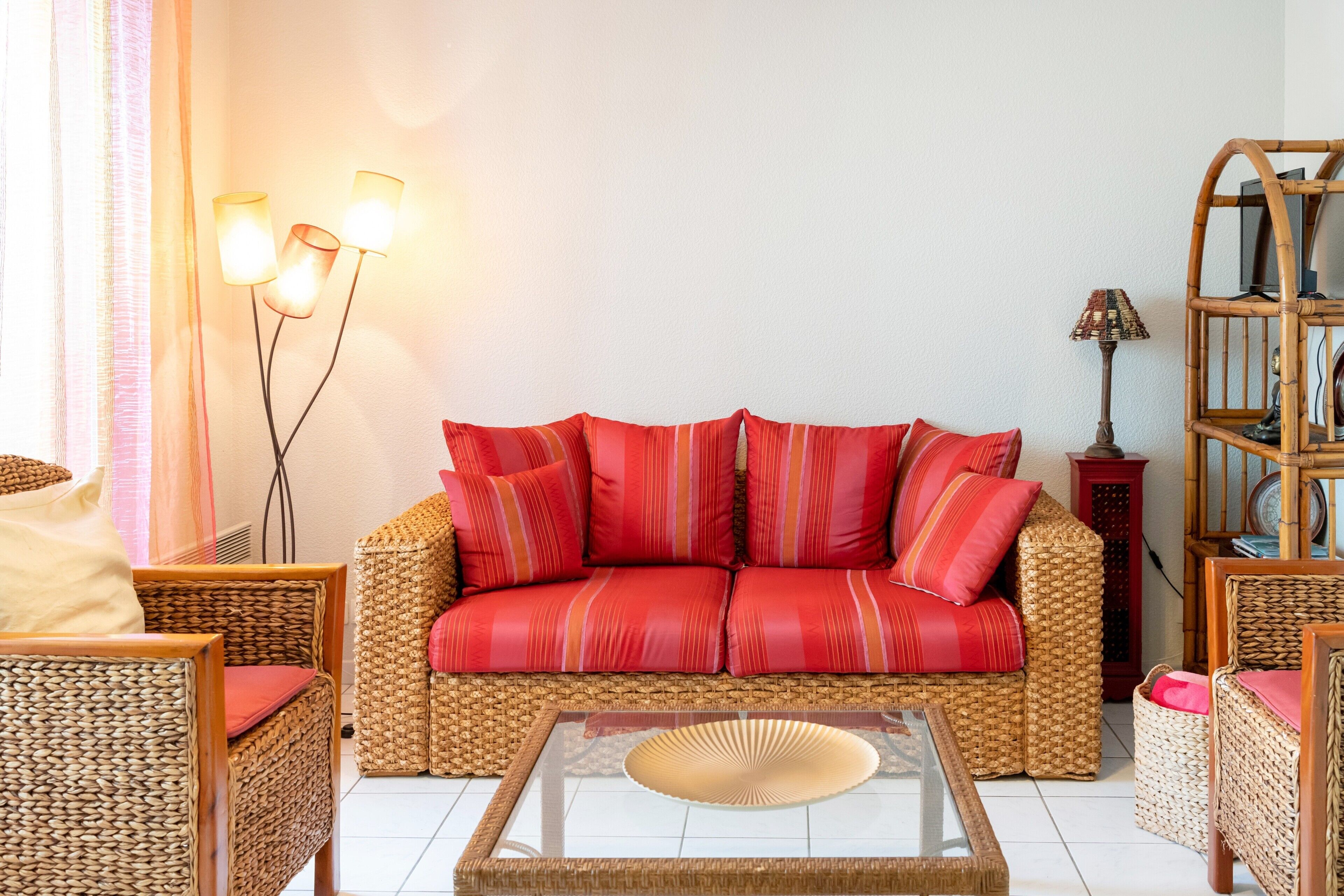 A wicker sofa with red cushions, a glass coffee table, wicker chairs, a lamp, and a bookshelf.