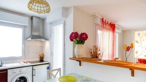 Una cocina con lavadora, una mesa de comedor con individuales, una lámpara colgante y una ventana con cortinas.