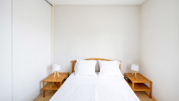 Una cama individual con ropa de cama blanca en una habitación con paredes lisas y dos mesitas de noche de madera con lámparas.