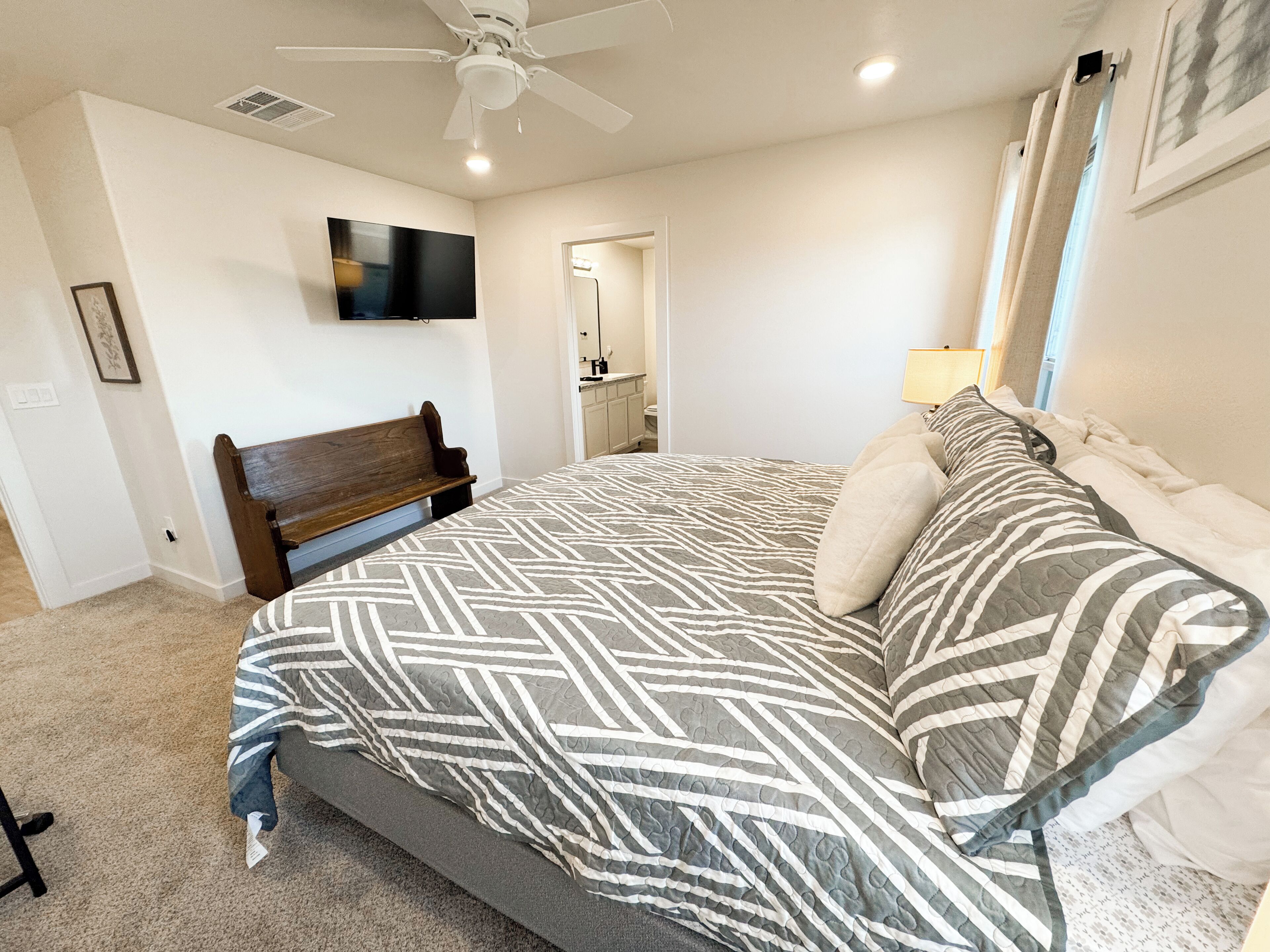 Une chambre à coucher comprenant un lit, un banc, un ventilateur de plafond, un téléviseur et une salle de bain visible à travers une porte ouverte.
