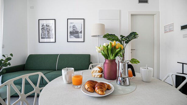 Uma mesa de jantar com croissants, suco de laranja, um vaso de flores e uma cafeteira.