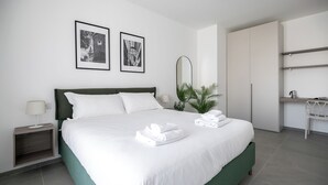 Quarto de hotel moderno com uma cama grande, mesa de cabeceira, espelho, planta e escrivaninha com cadeira.