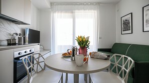 A modern kitchen with a dining table, chairs, a green sofa, and a window with curtains. - Italianway Comfort - Pasolini 8/G A (Milano)