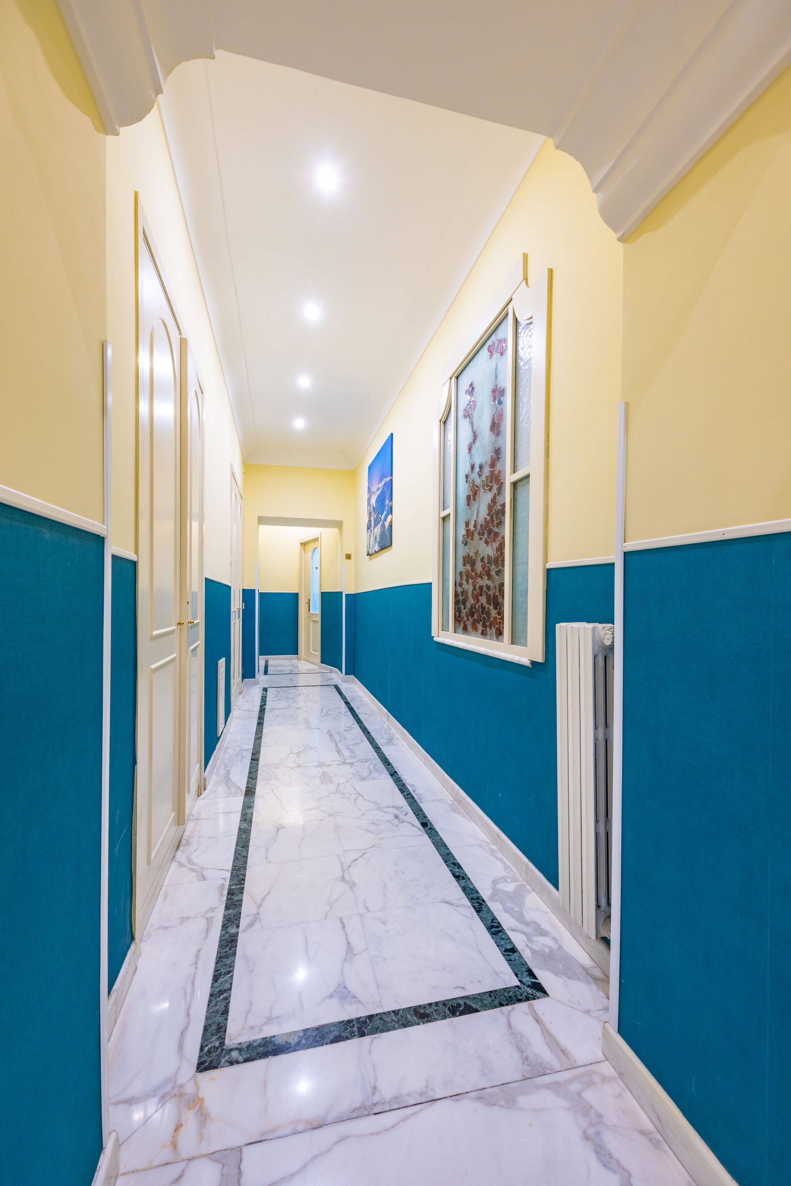 Un pasillo con suelo de mármol, paredes azules y una puerta blanca a la izquierda.