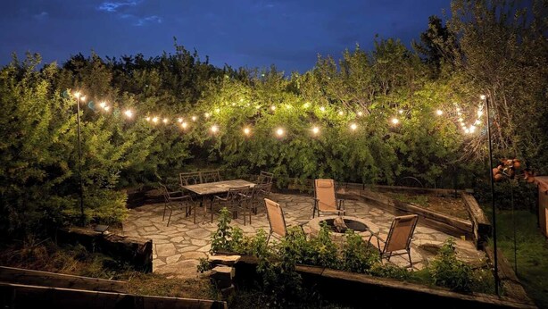 An outdoor patio with stone flooring, wooden chairs, a fire pit, and string lights.