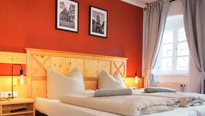A bedroom with a wooden headboard, a bed with white linens, two framed pictures on the wall, and a window with curtains.