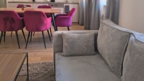 A modern living room with a sofa, a dining table with chairs, a kitchen area, and a window with curtains.