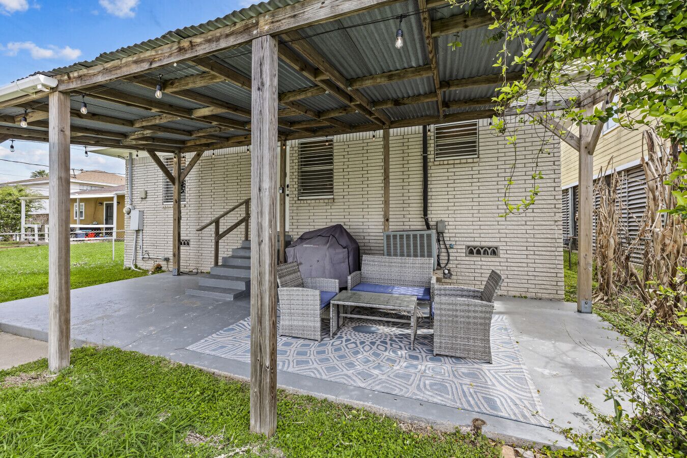 Een overdekt terras met tuinmeubilair, een barbecue en een bakstenen gebouw op de achtergrond.