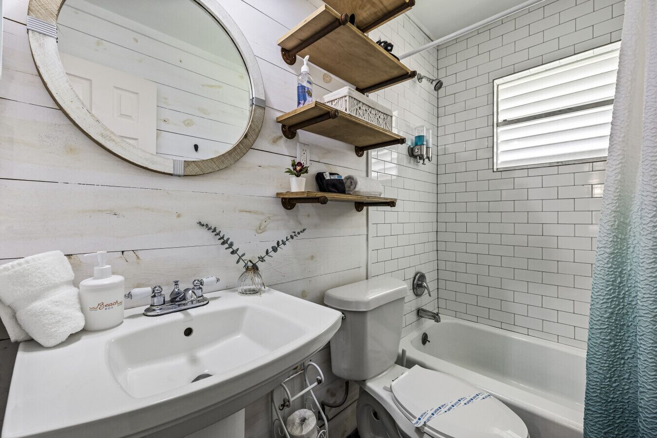 A bathroom with a white sink, toilet, and bathtub, wooden shelves above the sink, and a mirror on the wall.