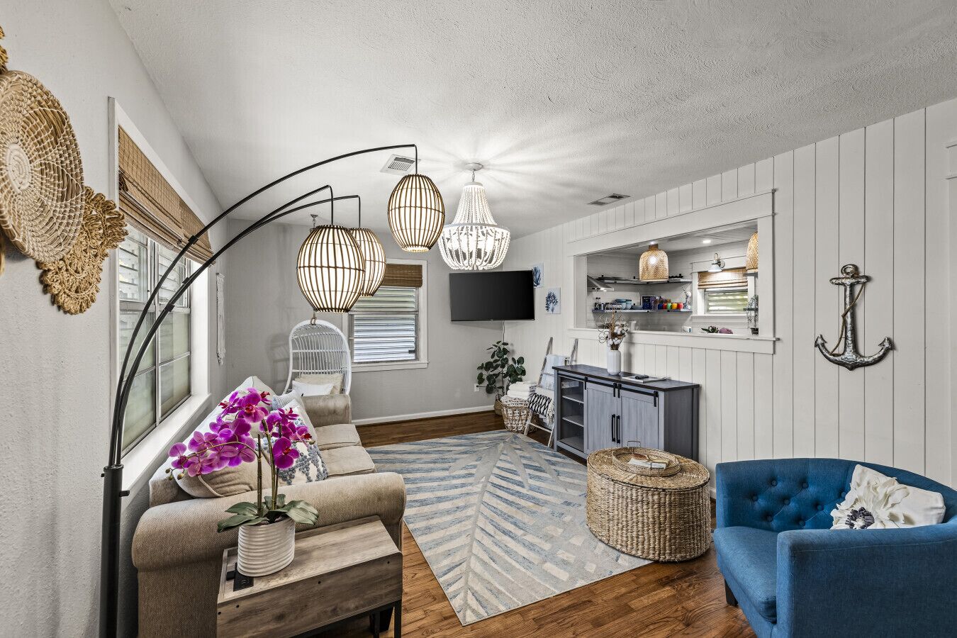 Un salon comprenant un canapé, un fauteuil bleu, un téléviseur, un coin repas et une cuisine avec un luminaire suspendu.
