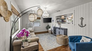 A living room with a sofa, a blue armchair, a TV, a dining area, and a kitchen with a hanging light fixture.