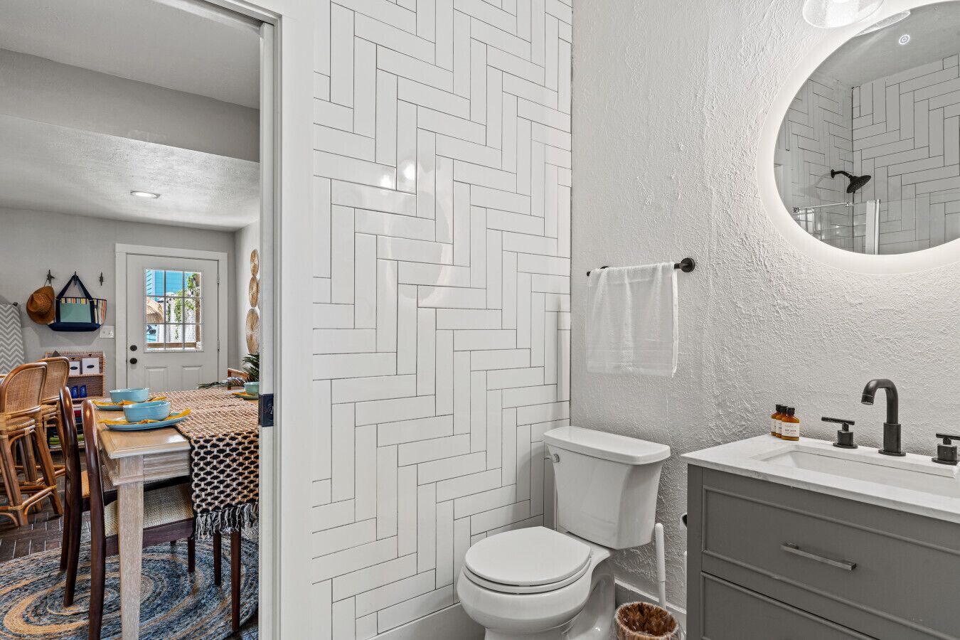 A bathroom with a white herringbone tile wall, a round mirror, a white toilet, and a grey vanity with a sink and faucet.