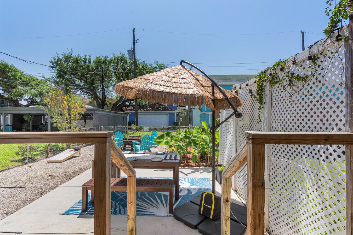 Un jardin avec une terrasse en bois, un parasol en chaume, du mobilier d’extérieur et une clôture blanche en treillis.