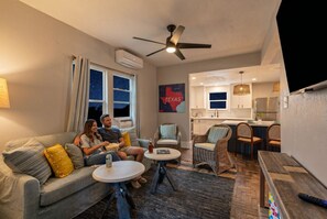 Ein gemütliches Wohnzimmer mit einer Couch, zwei Beistelltischen, Korbstühlen, einem Deckenventilator und einer Küchenzeile mit Esstisch und Stühlen.