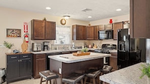 A modern kitchen with dark wood cabinets, a central island with stools, a black refrigerator, and a microwave. - Poconos Fun by Avantstay Mount Pocono Escape Firepit + Game Room Near Trails + Lakes (Long Pond)